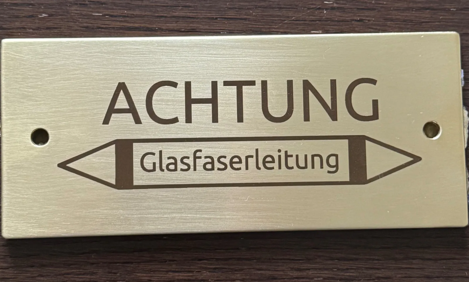 Graviertes messingfarbenes Hinweisschild mit der Aufschrift »Achtung Glasfaserleitung« und Warnpfeil